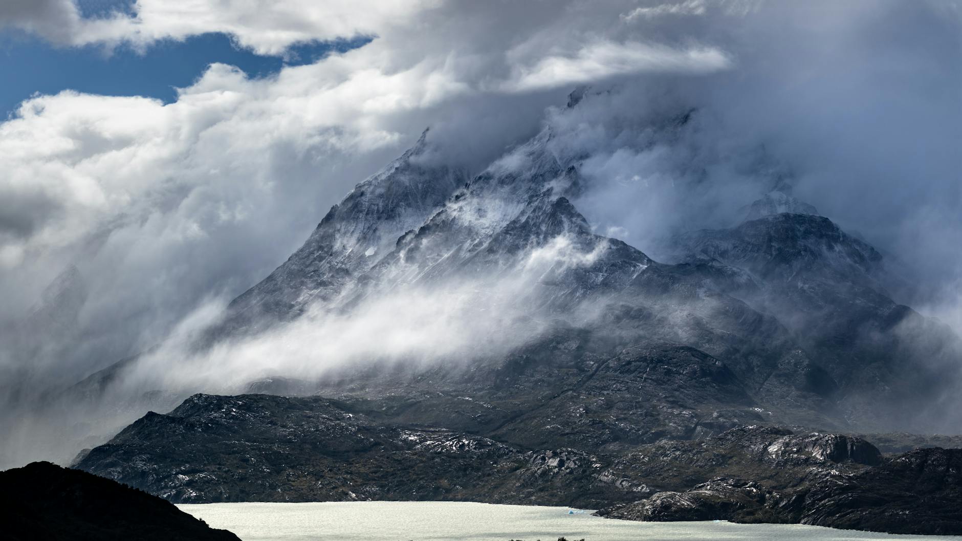 Dramatic mountain landscape under cloudy skies with mist and a body of water in the foreground.